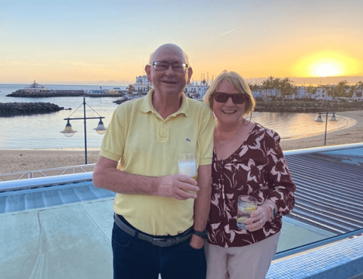 Two people holding drinks and standing on a rooftop overlooking a sandy beach at sunset 