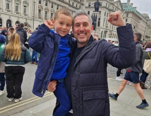 A man carrying his son on a busy street in London, throwing their fists in the air in happiness 