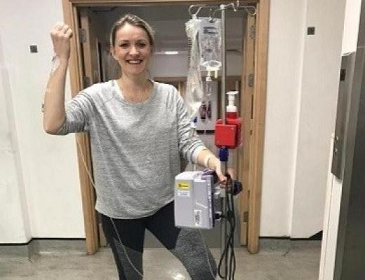 A woman smiling and standing in a Royal Marsden hospital corridor hooked up to a drip on wheels, through a cannula in her arm