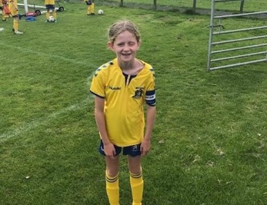 A young girl standing and smiling on a football pitch, wearing a bright yellow football kit with a ball by her feet