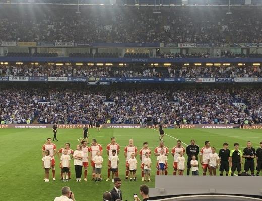 A football ground full of fans, with a line of players on the pitch and child mascots in front of them wearing RMCC t-shirts