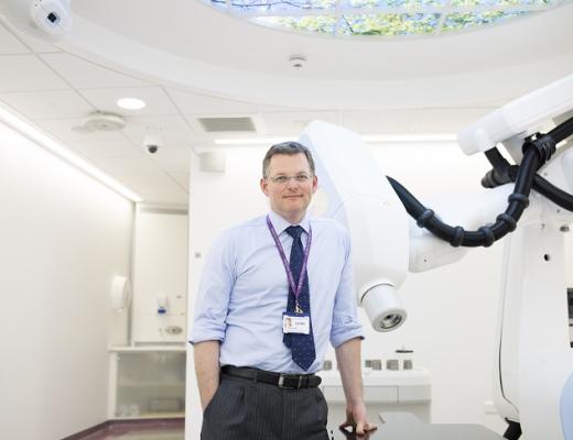 A smartly-dressed Royal Marsden doctor with a lanyard, standing next to a large white machine with a robotic arm