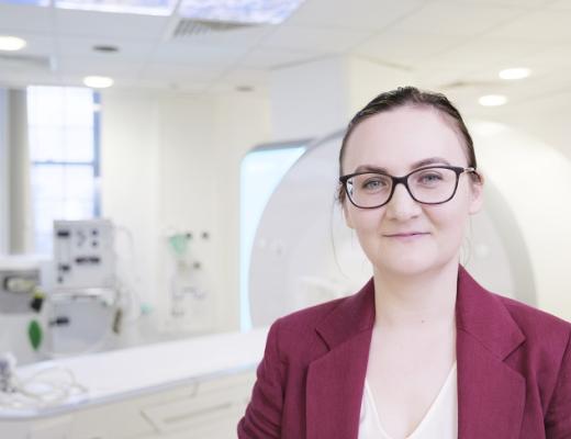 A doctor dressed smartly in a dark pink suit, wearing glasses and hair tied back, standing in front of a white machine like looks like an MRI