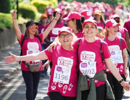 Two ladies wearing pink t-shirts, caps, and fundraising event numbers smile at the finish line of the Banham Marsden March