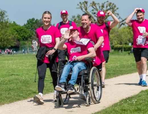 A group of six fundraisers wearing bright pink Banham Marsden March t-shirts. They are smiling and walking along a path together. One of the fundraisers at the front is smiling and waving at the camera - he is in a wheelchair. 