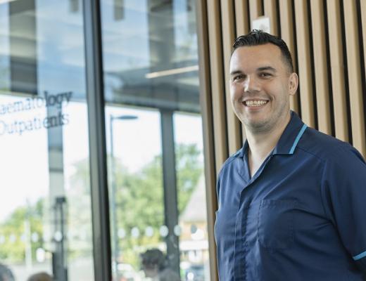 A man in a blue nurse outfit smiling in front of the modern Oak Cancer Centre building with wood panelling and glass