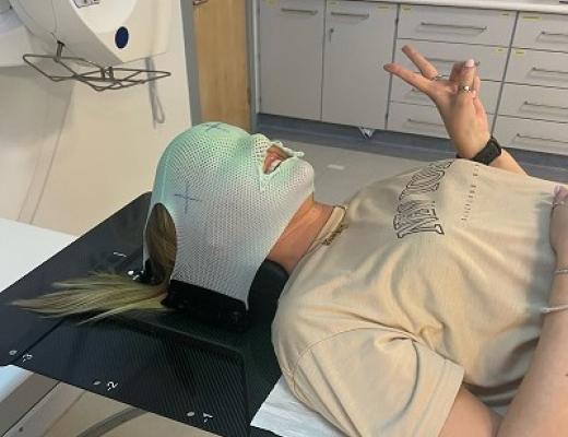 a person lying on a table in a hospital room, with a mask over her face and head. She's smiling and giving a peace sign