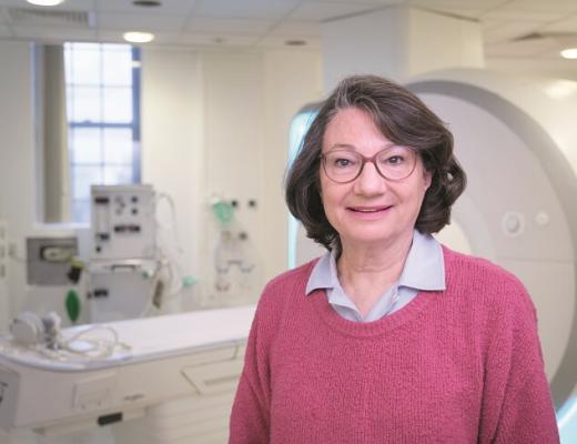 A Royal Marsden patient standing in front of an MRI machine in a hospital room, wearing a bright pink jumper and pink glasses