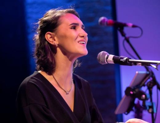 A young person dressed formally on a stage smiling and singing into a microphone 