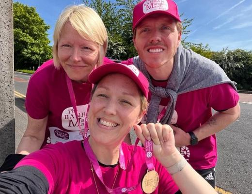 A Royal Marsden patient in a wheelchair smiling and holding up a Banham Marsden March medal, with two people smiling behind her.