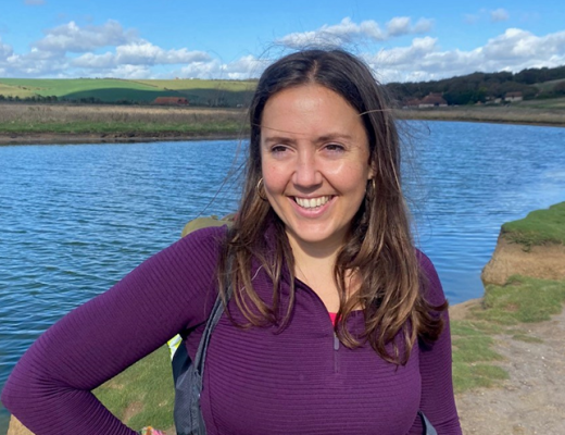 Tabitha smiling, wearing purple hoodie and standing in front of a lake