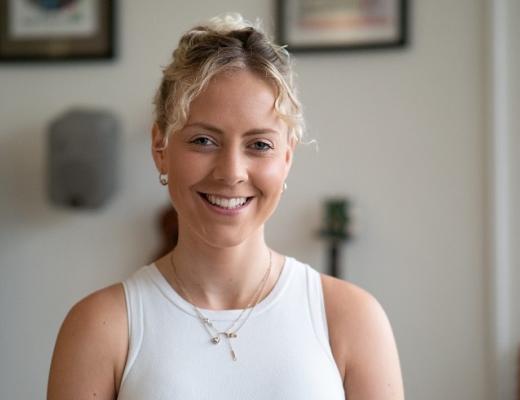 The head and shoulders of a young woman smiling, wearing her hair tied up and a white summer top with gold jewellery