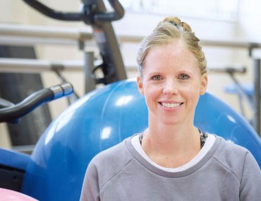 A Royal Marsden Physiotherapist smiling, wearing a sports jumper and sitting in front of gym equipment including a yoga ball and exercise bike.