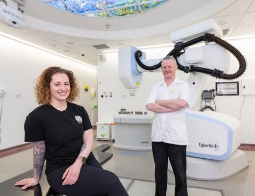 A smiling woman sitting in a hospital treatment room, beside a man dressed in hospital staff uniform. In the background there is radiotherapy equipment.  