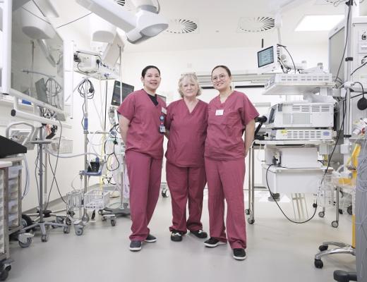 Three people in pink hospital scrubs, standing in a hospital room filled with medical equipment