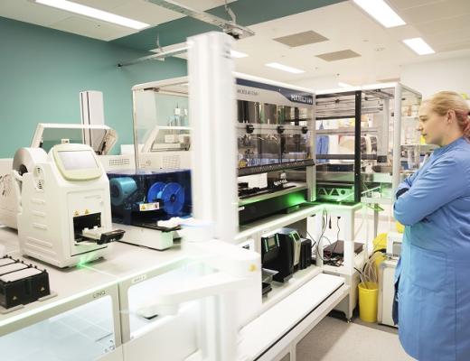 A large white robot with a moving arm picking up slides, in a Royal Marsden laboratory. It is being watched by a member of the lab team.