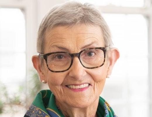 Head and shoulders of a woman with short grey hair, she's smiling and wearing stylish glasses and dark blue and green scarf