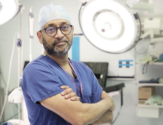 A surgeon at The Royal Marsden, standing in an operating theatre with equipment behind him. He has is arms crossed and is wearing blue scrubs