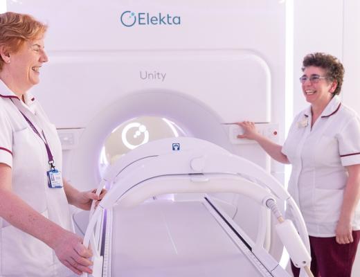 Two radiographers from The Royal Marsden smiling and laughing. They're standing next to a large white machine that looks like an MRI scanner.