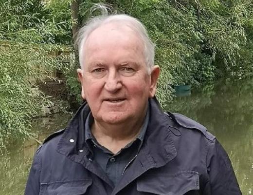 Head and shoulders of Phillip, he has short, silver hair and is wearing a black jacket. He's outside, standing in front of a green, leafy pond.