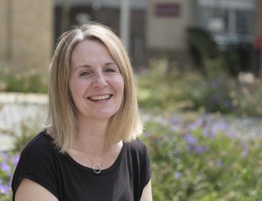 A Royal Marsden patient sitting in the flowery front garden of the Oak Cancer Centre. She has short blonde hair and a black top, she's smiling.