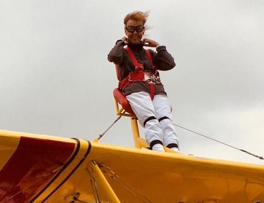 Marilyn on the wing of the plane, she is strapped in with a safety harness and wearing goggles. Her hair is blowing in the wind