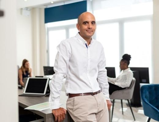 A man in white shirt and beige trousers, leaning against an office desk
