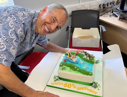 Dennis wearing a blue, pattered shirt. He's smiling and leaning over a 60th birthday cake that is decorated with icing figures of him and his dog canoeing down a river.