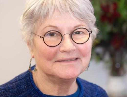 A smiling headshot of Royal Marsden patient, Betsy. She has short, cropped silver hair and round, dark-framed glasses. She's wearing a dark blue knitted jumper and some dangly earrings. 