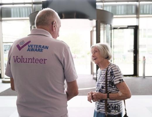 Brain in a light pink polar neck shirt that reads 'Veteran aware volunteer' on the back. He's speaking to a patient in the entrance of The Royal Marsden's Oak Cancer Centre