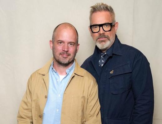 Jack Dyson and Daniel Marks, founders of father and son day. Jack is sitting down and wearing a light blue shirt with a cream-coloured jacket over the top. Daniel is standing next to him, wearing dark framed glasses and a navy blue jacket. 