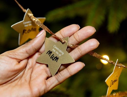 A close up photo of a hand holding a golden star decoration, hung on a christmas tree. The star says 'Celebrating the life of Mum' with the word 'mum' handwritten on the star.