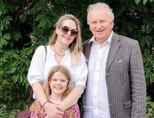 Philip standing outside in front of some green foliage with his daughter and granddaughter. 